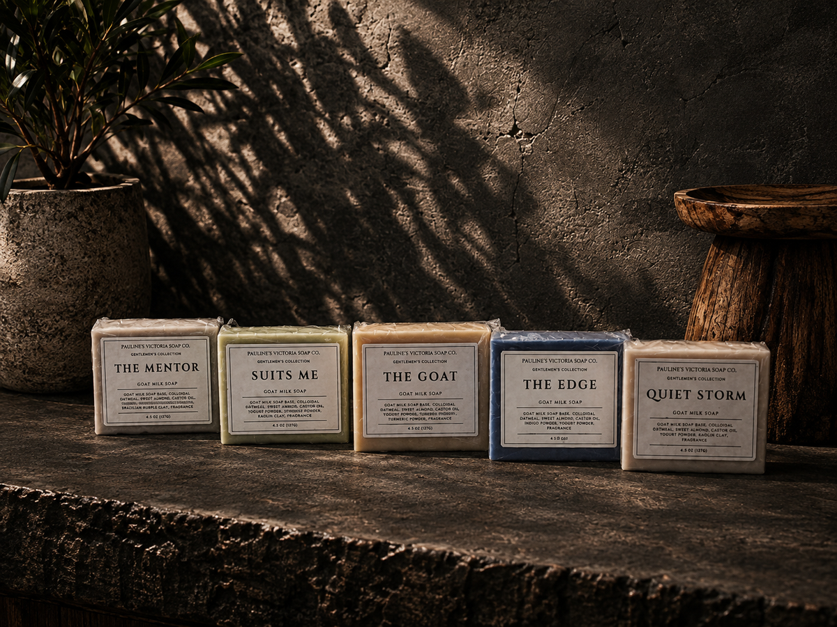 Five bars of soap in different colors lined up on a rustic stone surface against a textured wall with leaf shadows. The soaps are labeled 'THE MENTOR', 'SUITS ME', 'THE GOAT', 'THE EDGE', and 'QUIET STORM'.