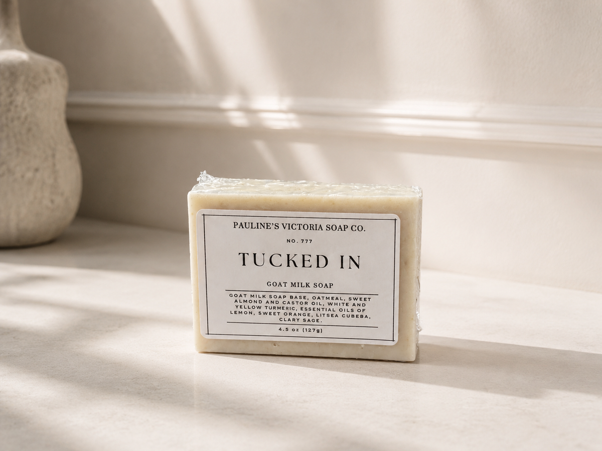 A rectangular bar of soap labeled 'Tucked In' by Pauline's Victoria Soap Co., placed on a light stone surface with soft window light and shadows.